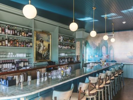 A stylish bar interior with a long marble counter, teal accents, hanging globe lights, shelves of bottles, and mint-green seating along the bar.