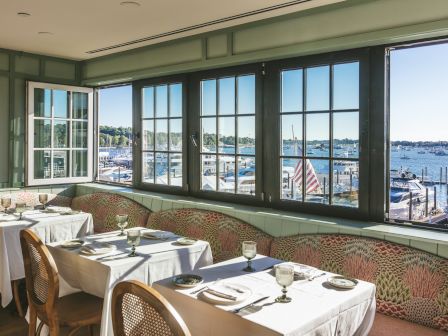 A seaside restaurant with large windows, white-draped tables, woven chairs, and a harbor view with boats outside, serene and elegant.