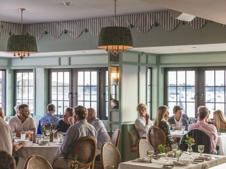 A bustling restaurant dining room with patrons at tables, natural light from large windows, wicker chairs, and green striped decor, lively ambiance.