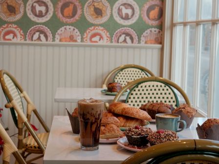 A cozy cafe scene with a small table of pastries, coffee, and drinks, rattan chairs, and retro wallpaper by a sunny window.