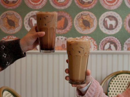 Two people clink iced coffees in a cozy cafe, patterned wallpaper in the background, both hands visible, warm and casual vibe.