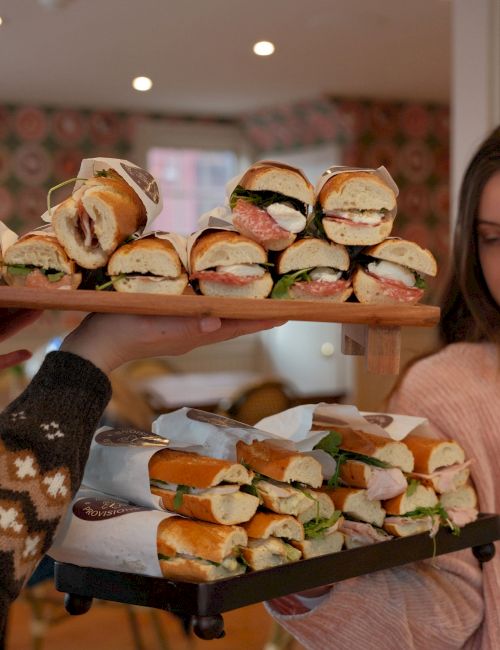 A tray of mini sandwiches with cured meats, greens, and cheese is being held at a cafe.
