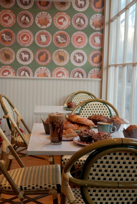 A sunlit breakfast table with pastries, croissants, coffee, and juice, set inside a cozy room decorated with colorful bird-pattern plates on the wall.